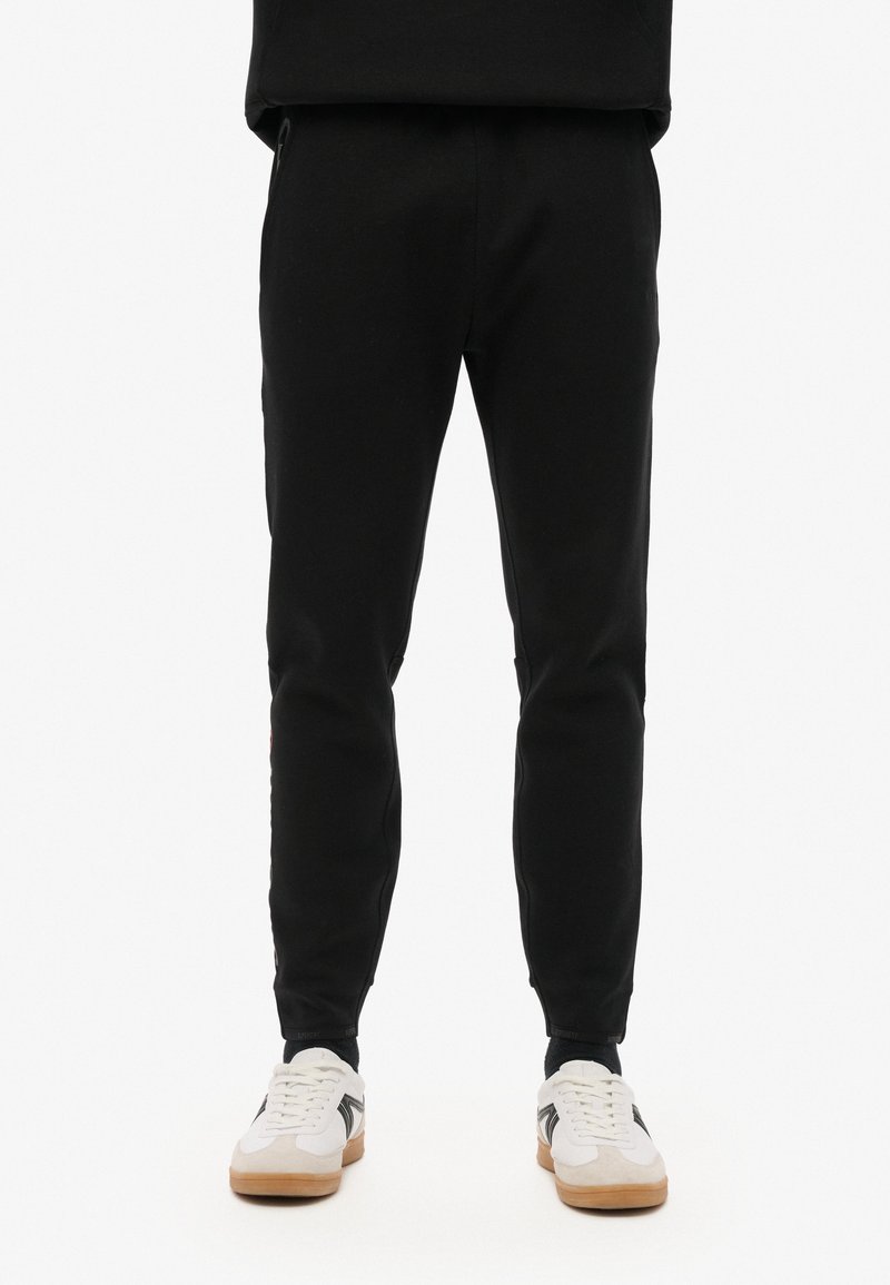 Zwarte joggers met een getailleerde pasvorm, uitgerust met twee zijzakken en een rood logodetail, gecombineerd met witte sneakers.