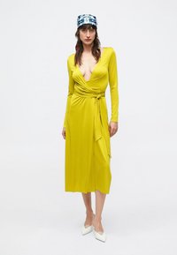 Rochie cu mâneci lungi, de tip petrecut, din țesătură galbenă strălucitoare. Are un decolteu adânc în V, detaliu cu legătură laterală și fustă lungime midi.