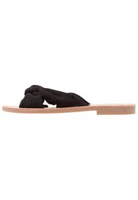 Black suede slide sandal with knotted upper, flat tan sole, and minimalistic design. Features a smooth texture and no embellishments.