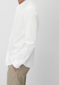 Vit bomullsskjorta med ståkrage, knäppning fram och långa ärmar. Texturerad tyg, avslappnad passform, bärs med ljusa khakibukser.