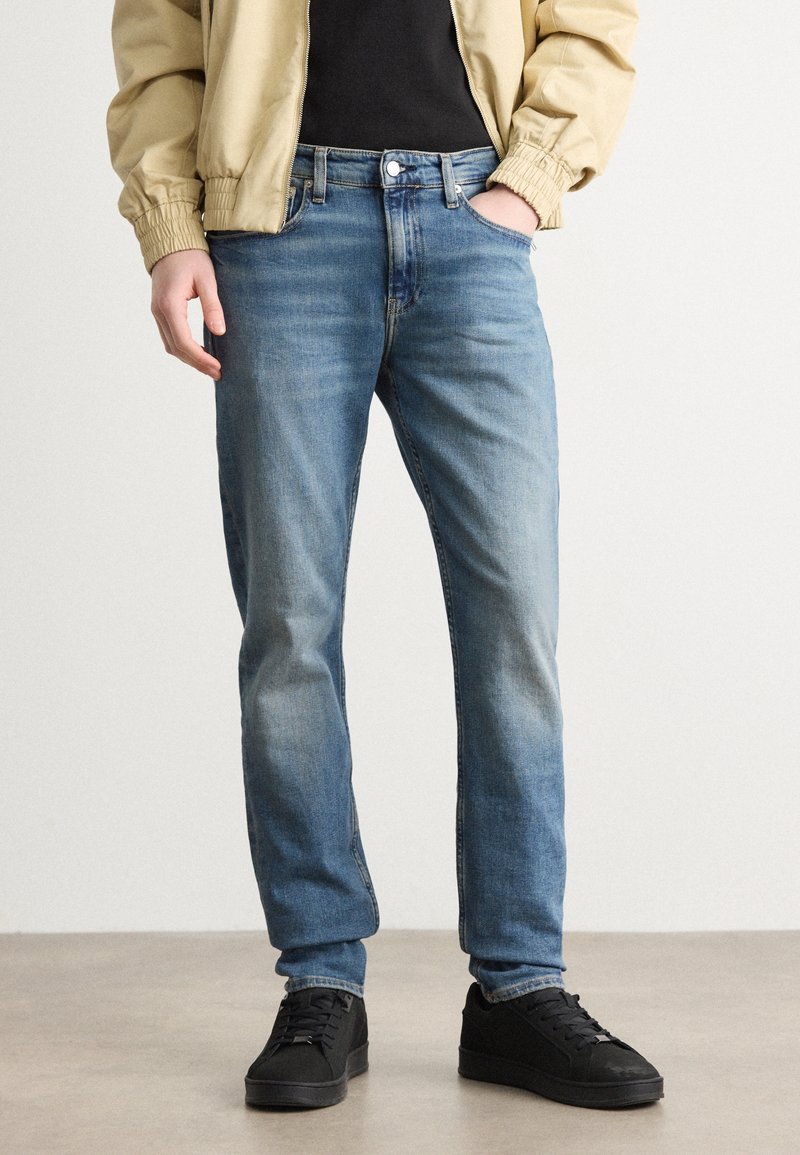Personne portant un jean slim bleu clair, des baskets noires, une chemise noire et une veste beige avec la main dans la poche, debout sur un sol gris.