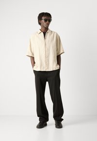 Lichtbeige kortemouwen shirt met knopen, zwarte broek en zwarte sneakers. Model draagt zwarte zonnebril; soepele textuur.