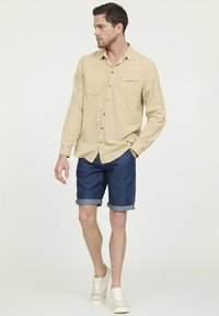 Beige overhemd met lange mouwen en knoopsluiting aan de voorkant, voorzien van één zak, gedragen met opgerolde denimshorts en lichte sneakers, tegen een neutrale achtergrond.