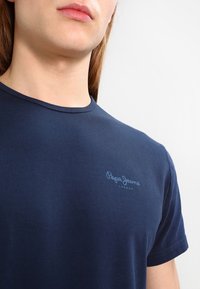 Młoda osoba ubrana w granatową koszulkę Pepe Jeans London, przycięta od szyi do górnej części torsu, na jednolitym białym tle.
