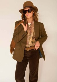Blazer marron ajusté sur une chemise à motifs, associé à un pantalon en velours côtelé noir. Les accessoires comprennent un chapeau en feutre marron, des lunettes de soleil et un sac à franges.