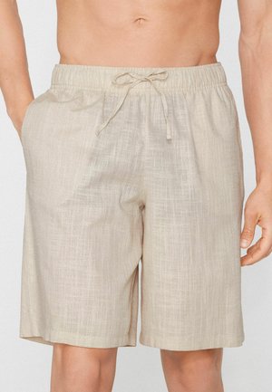 Beige linnen shorts met trekkoord, gedragen door een persoon met handen deels in de zakken, waarbij de buik en bovenbenen zichtbaar zijn.