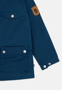 Dark blue jacket with a textured finish, featuring a tan leather patch, snap buttons, and a front pocket. Accents include a small flag label.