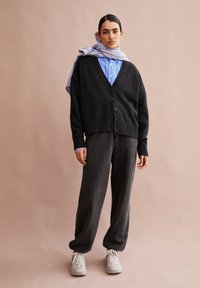 Zwarte wollen cardigan met knoopsluiting, gelaagd over een blauw gestreept shirt. Donkergrijze sweatpants en lichtgrijze sneakers maken de outfit compleet.