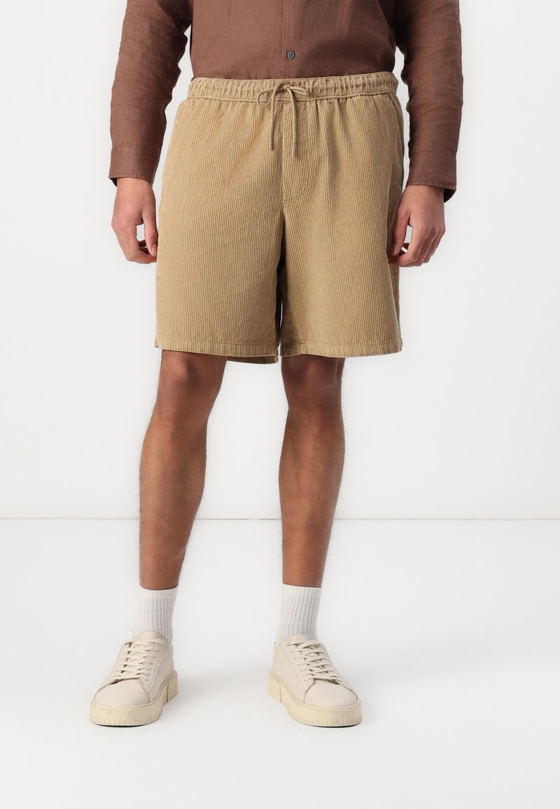 Shorts en velours côtelé beige avec taille élastique et cordon de serrage. Présente une coupe décontractée et une texture ridée verticale. Portés avec des baskets blanches.