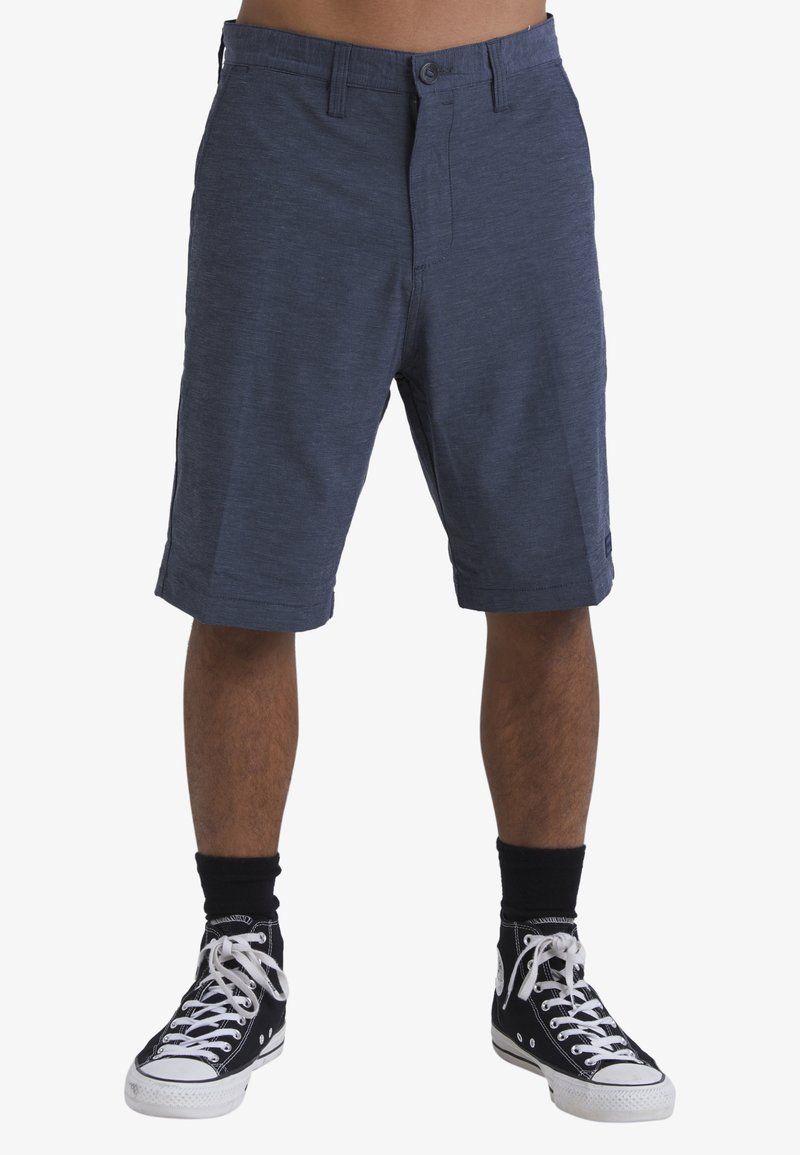 Uomo che indossa pantaloncini blu navy fino al ginocchio, sneaker alte nere con lacci bianchi e calzini neri, in piedi davanti a uno sfondo semplice.