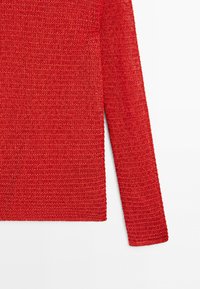 Close-up of a textured red knit sweater sleeve and side against a white background, showing detailed stitch pattern.