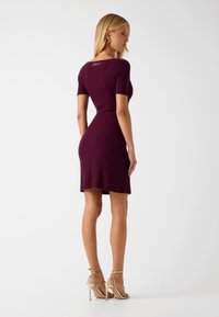 Knielange, geribbelde jurk in donker bordeauxrood met korte mouwen en een aansluitende pasvorm. Gecombineerd met gouden sandaalhakken.