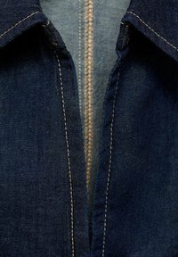 Jeanshemd in einem tiefen Blau mit spitzem Kragen und kontrastierendem hellblauem Innenstoff. Sichtbare Nähte in goldener Farbe.