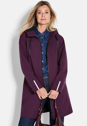 Femme portant un manteau à capuche violet par-dessus une chemise en jean et un jean, tenant un sac à main violet assorti devant un fond clair uni.
