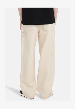 Beżowe spodnie cargo o luźnym kroju, wykonane z trwałego materiału. Posiadają liczne kieszenie oraz widoczne szwy. U dołu widoczne czarne trampki.