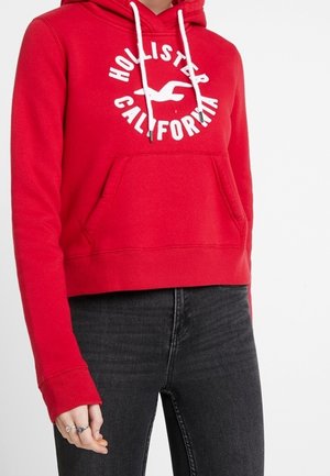 Czerwona bluza z kapturem Hollister California z białym logo i sznurkami, noszona z czarnymi jeansami, pokazująca częściowy widok osoby.