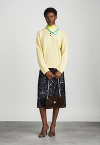 Keltainen neulepaita, jossa on kolme nappia, yhdistettynä tummansiniseen paljettihameeseen. Pitelee ruskeaa mokkanahkakäsilaukkua, jossa on ketjuhihna. Kirkkaat korkokengät.