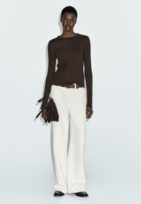 Maglietta marrone a maniche lunghe abbinata a pantaloni bianchi a gamba larga. La modella tiene una piccola borsa in suede nera. Scarpe nere basse completano l'outfit.
