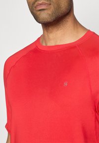 Κόκκινο αθλητικό t-shirt με κοντά μανίκια, στρογγυλή λαιμόκοψη και διακριτική λεπτομέρεια λογότυπου. Φτιαγμένο από απαλό, ελαφρύ ύφασμα με εφαρμοστή γραμμή.