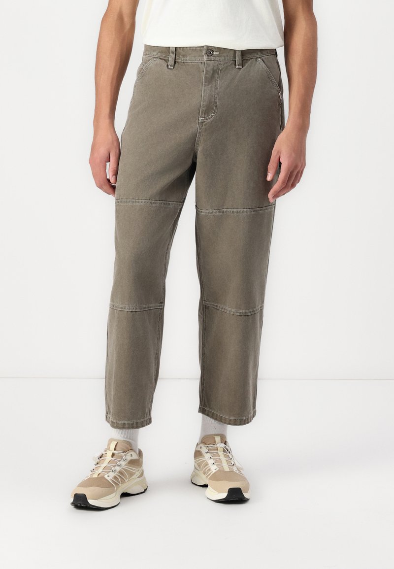Un pantalon en coton kaki présente une coupe droite avec des coutures horizontales et des détails de surpiqûres discrets, associé à des chaussures de sport beiges.