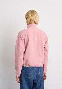 Persoon met kort blond haar, gekleed in een lichtroze fleece jas en blauwe jeans, kijkend naar een eenvoudige witte muur.