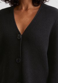 Schwarzer Strickcardigan mit V-Ausschnitt, ausgestattet mit vier großen Knöpfen und einer strukturierten Oberfläche. Der Stoff ist weich und hat eine leichte Beschaffenheit.