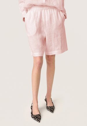 Mujer con pantalones cortos de lino rosa claro y tacones slingback puntiagudos con estampado de cebra en negro y beige, de pie contra un fondo claro.