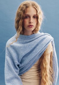 Hellblauer Pullover aus weichem Material, lässig über die Schultern drapiert; mit einem gerippten Ausschnitt und langen, welligen Haaren im Vordergrund.
