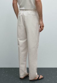 Pantaloni in cotone beige chiaro con una vestibilità comoda, cintura elastica e tasche posteriori, abbinati a infradito nere, visti da dietro.