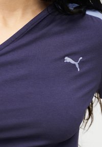 Κοντινό πλάνο ενός ατόμου που φοράει μωβ μπλούζα με V-λαιμόκοψη και λευκό λογότυπο Puma στο αριστερό μέρος του στήθους.