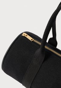 Μαύρη κυλινδρική τσάντα με υφασμάτινες λαβές υφής και χρυσό φερμουάρ σε λευκό φόντο.