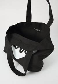 Sac fourre-tout en toile noire, avec un sommet ouvert, deux poignées solides et un détail graphique blanc sur le devant. L'intérieur est non doublé.