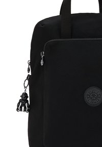 Sac à dos en tissu noir avec une surface texturée, doté d'une poche zippée à l'avant, d'un patch logo rond et d'un petit porte-clés en forme de singe noir attaché.
