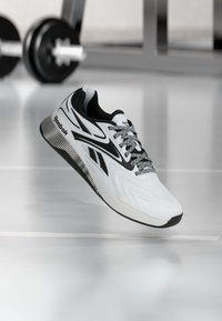 Άσπρο και μαύρο αθλητικό παπούτσι Reebok με κορδόνια με σχέδια που αιωρείται πάνω από το πάτωμα γυμναστηρίου, με βάρη γυμναστηρίου θολά στο φόντο.