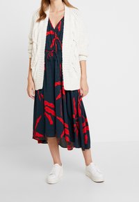 Bílý pletený kardigan s texturovanými drobnými vzory, přehozený přes tmavé šaty s červenými abstraktními vzory, doplněný bílými teniskami.