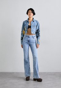 Iškirpta mėlyna džinsinė striukė su juodais gėlių raštais, derinama su aukštu liemeniu šviesiai mėlynomis džinsinėmis kelnėmis ir rudomis platforminėmis bateliais.