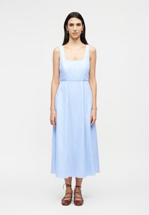 Γυναίκα με μακριά σκούρα μαλλιά, ντυμένη με μίντι φόρεμα χωρίς μανίκια σε ανοιχτό μπλε και λευκές ρίγες, και καφέ παπούτσια με κορδόνια, στέκεται μπροστά σε απλό φόντο.