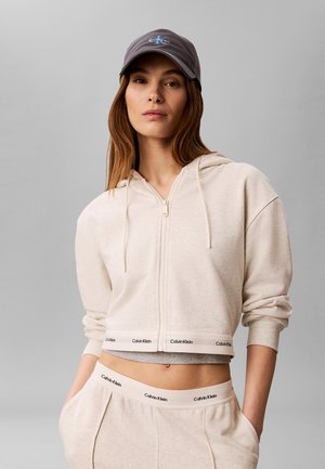 Kobieta w beżowej bluzie na zamek i dopasowanych spodniach z logo Calvin Klein na pasie oraz szarej czapce Calvin Klein, stojąca na szarym tle.