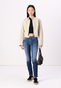 Chaqueta corta beige con cuello alzado, llevada sobre un top corto negro, combinada con vaqueros skinny azules y zapatillas negras, llevando un bolso oscuro.