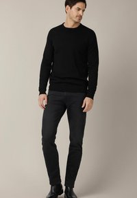 Czarny sweter z okrągłym dekoltem wykonany z dzianiny, zestawiony z ciemnoniebieskimi dżinsami. Modelka stoi pewnie, nosząc botki za kostkę. Neutralne tło.