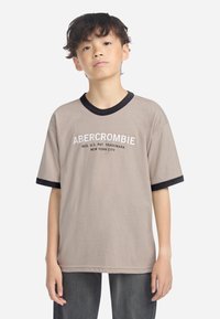 Beige t-skjorte med korte ermer som har svart ribbestrikket krage og mansjetter. "ABERCROMBIE" trykket i hvitt på brystet. Laget av bomull.