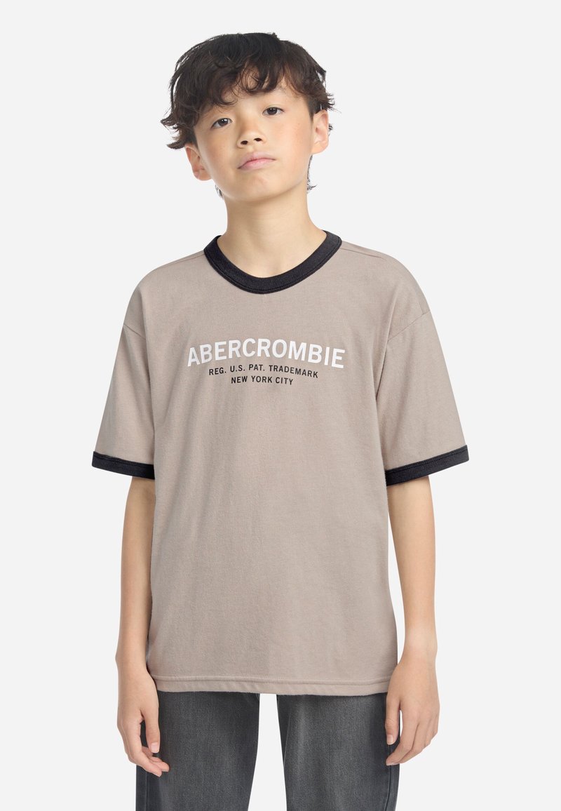 Beige t-skjorte med korte ermer som har svart ribbestrikket krage og mansjetter. "ABERCROMBIE" trykket i hvitt på brystet. Laget av bomull.