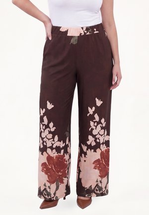 Kvinde iført brun viddebuks med store pink og røde blomster- og sommerfugleprint, kombineret med brune spidse hæle og en hvid top.