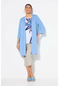 Hellblaues, langes Cardigan mit Dreiviertelärmeln über einem weißen Oberteil mit blauer Blumendekoration. Helle beige, cropped Hose und blaue Sandalen.