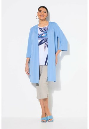 Cardigan long bleu clair à manches trois-quarts sur un haut blanc avec un motif floral bleu. Pantalons croppés beige clair et sandales bleues.