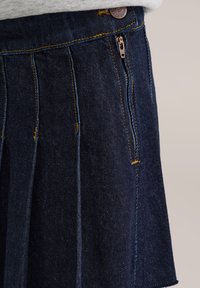 Temno moderna denim krila s pliseji, s kontrastnim rumenim šivom in zadrgo na strani. Tekstilirano blago in detajli s bakrenimi gumbi.