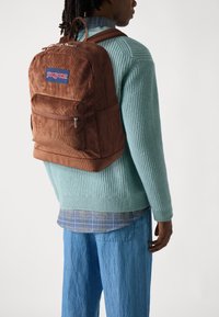 Καφέ σακίδιο από καπιτονέ ύφασμα με μπροστινή τσέπη που κλείνει με φερμουάρ και λογότυπο JanSport. Φοριέται πάνω από ένα ανοιχτό μπλε υφαντό πουλόβερ, συνδυασμένο με μπλε παντελόνι.