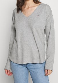 Lys grå langærmet v-hals top med en ribbet tekstur og diskret logo detalje på brystet. Rundet kant og afslappet pasform; parret med blå jeans.