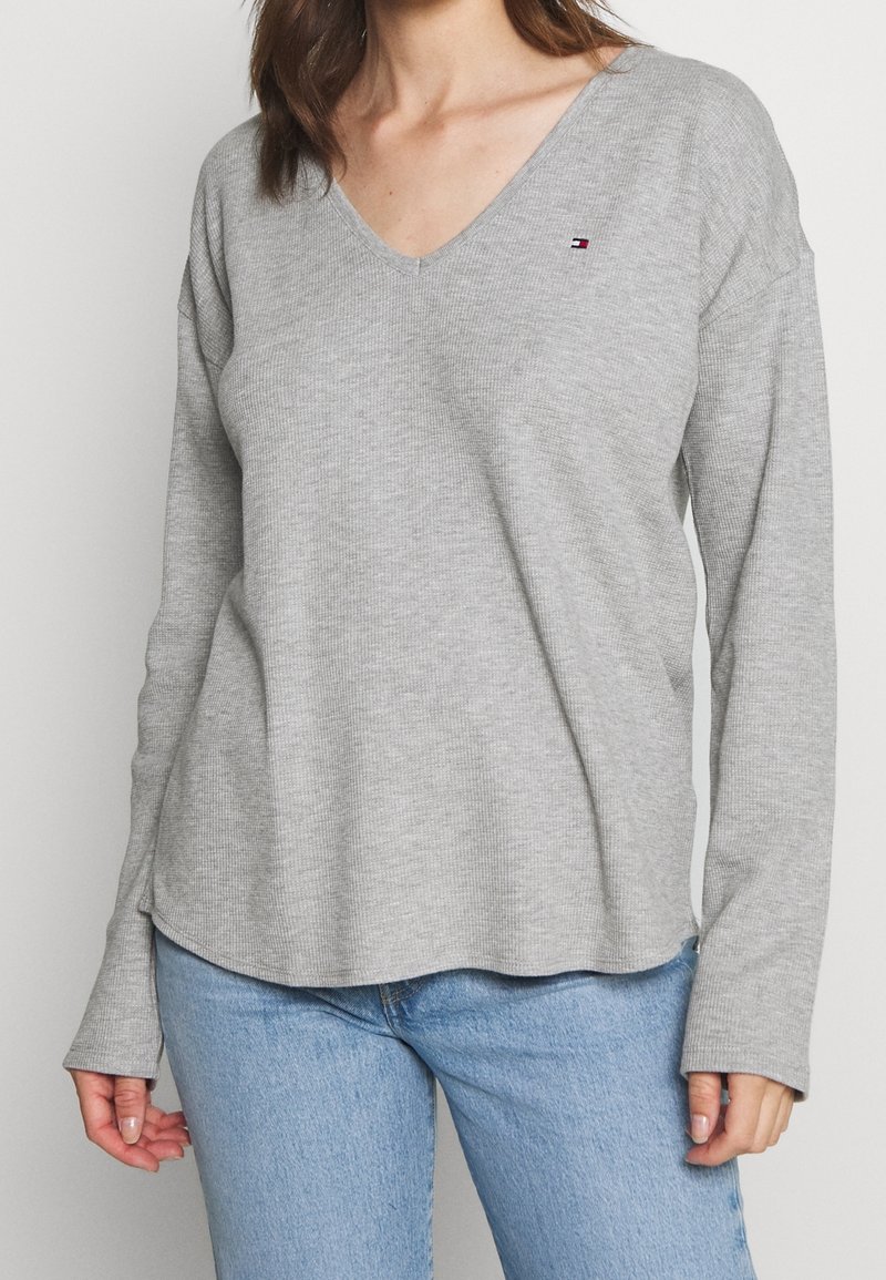 Lys grå langærmet v-hals top med en ribbet tekstur og diskret logo detalje på brystet. Rundet kant og afslappet pasform; parret med blå jeans.
