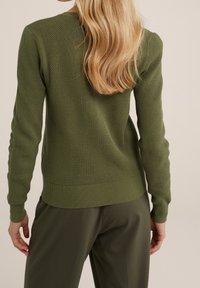 Olivově zelený pletený svetr s texturovaným diamantovým vzorem, s žebrovanými manžetami a lemem. Nosený s odpovídajícími olivově zelenými kalhotami.